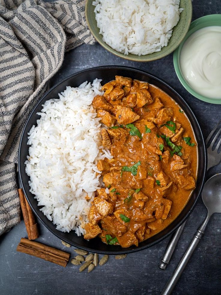 a black plate topped with rice and chicken curry next to a bowl of sour cream