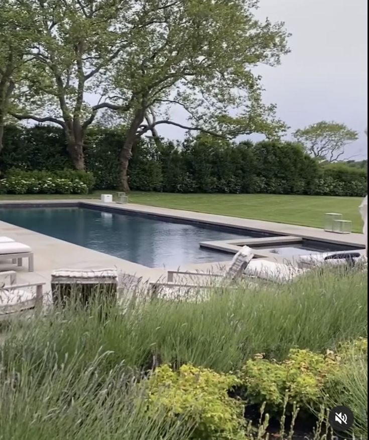 an empty swimming pool surrounded by tall grass and trees in the background, with chairs around it