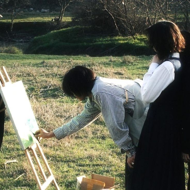 two women and a man are painting in the grass with one woman looking at an easel