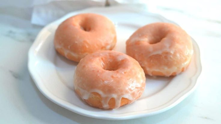 three glazed doughnuts on a white plate sitting on a marble counter top, ready to be eaten