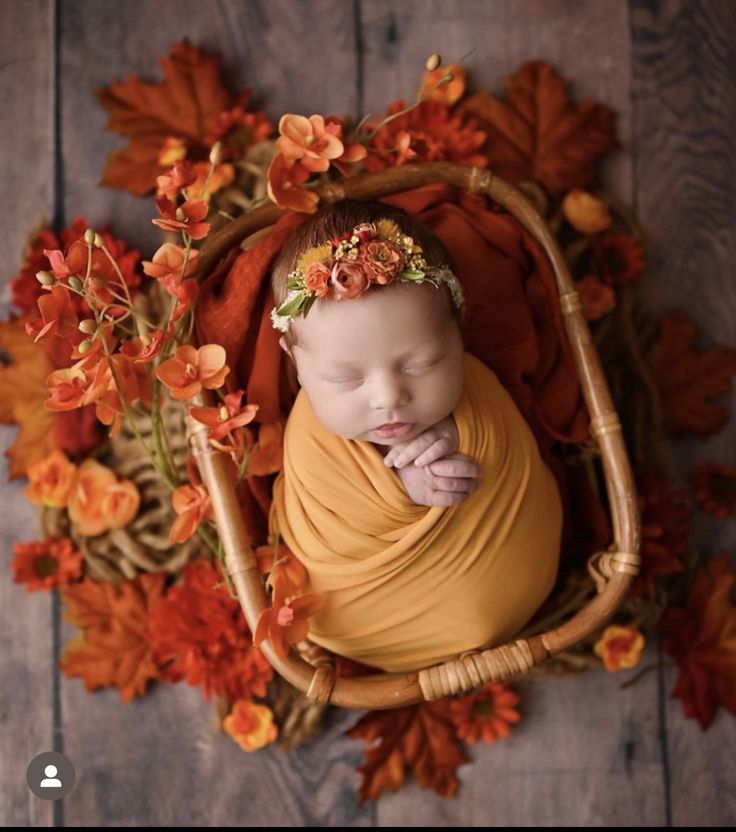 a newborn baby wrapped in a yellow wrap laying on a wooden floor surrounded by autumn leaves and flowers