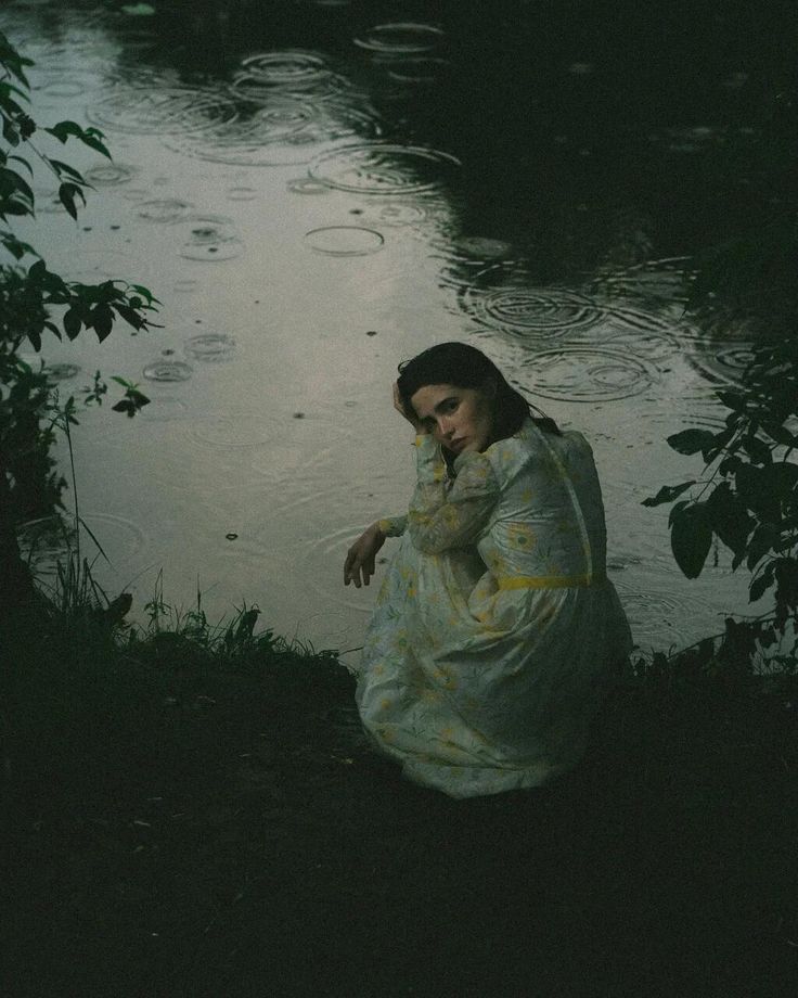 a woman sitting on the ground next to a body of water with raindrops