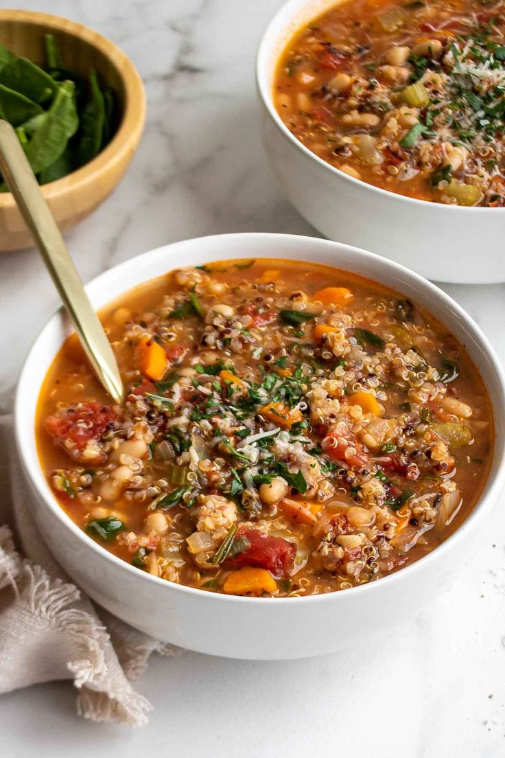 two white bowls filled with vegetable soup on top of a marble counter next to a bowl of spinach