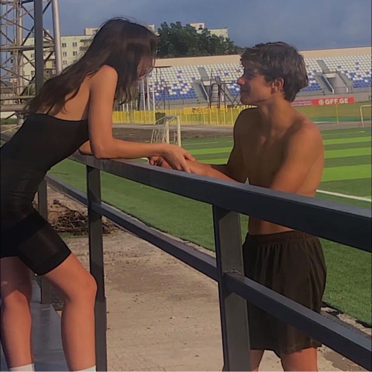 a young man and woman standing next to each other on a fence near a baseball field