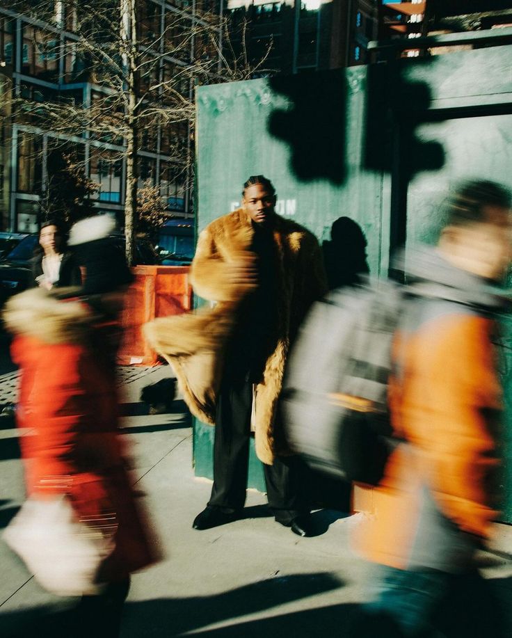 a man in a fur coat is standing on the sidewalk with other people walking by