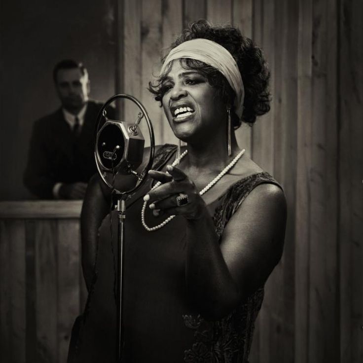 a black and white photo of a woman singing into a microphone with a man in the background