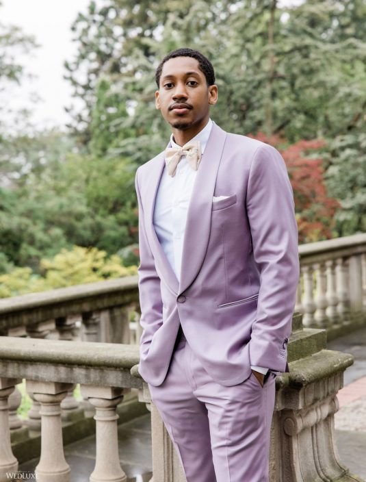 a man wearing a purple suit and bow tie standing next to a stone railing with trees in the background