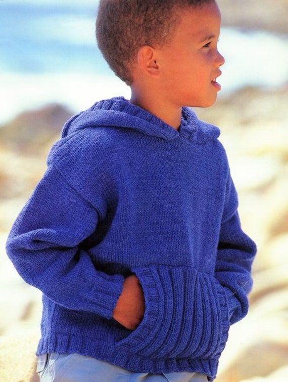 a young boy wearing a blue sweater standing on the beach with his hands in his pockets