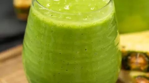 a green smoothie in a tall glass next to sliced pineapple on a cutting board