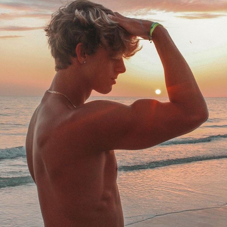 a shirtless man standing on top of a beach next to the ocean at sunset