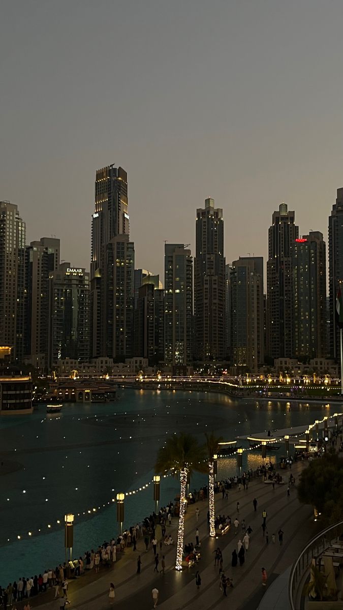 the city skyline is lit up at night and people are walking on the sidewalk near the water