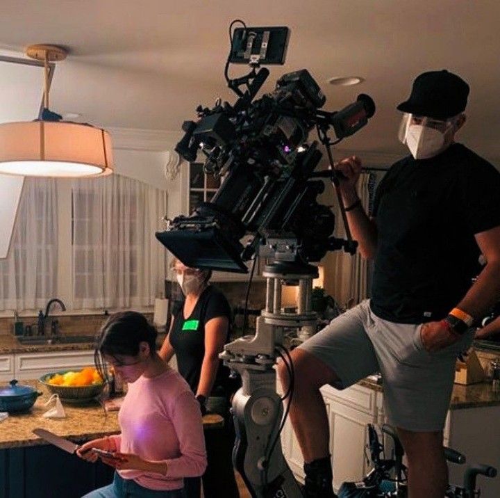 three people in a kitchen with cameras on the counter and one person wearing a face mask