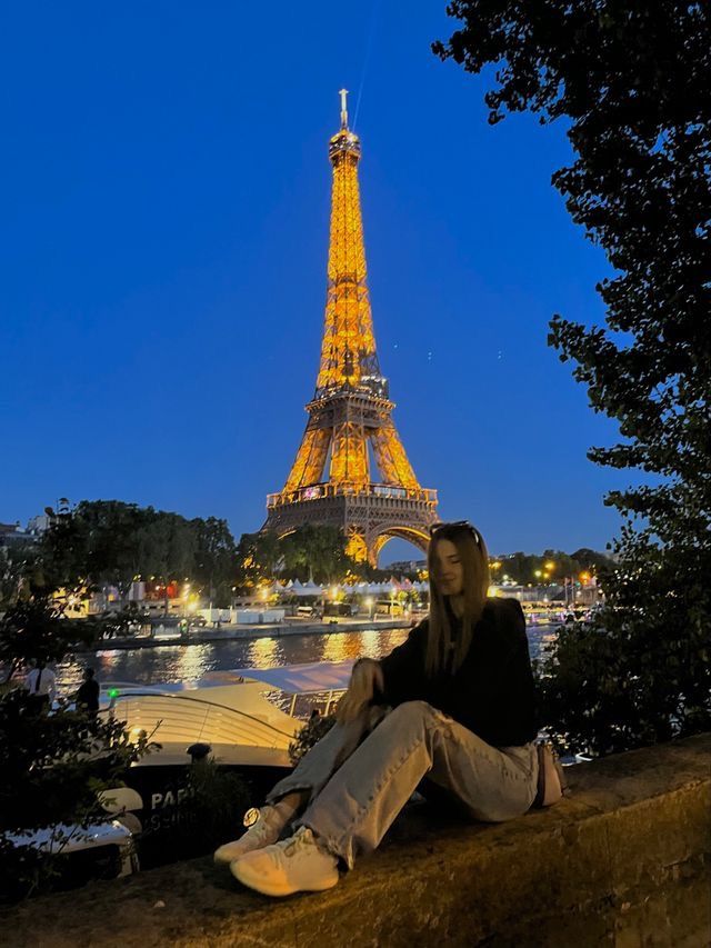 a woman sitting on a ledge in front of the eiffel tower at night