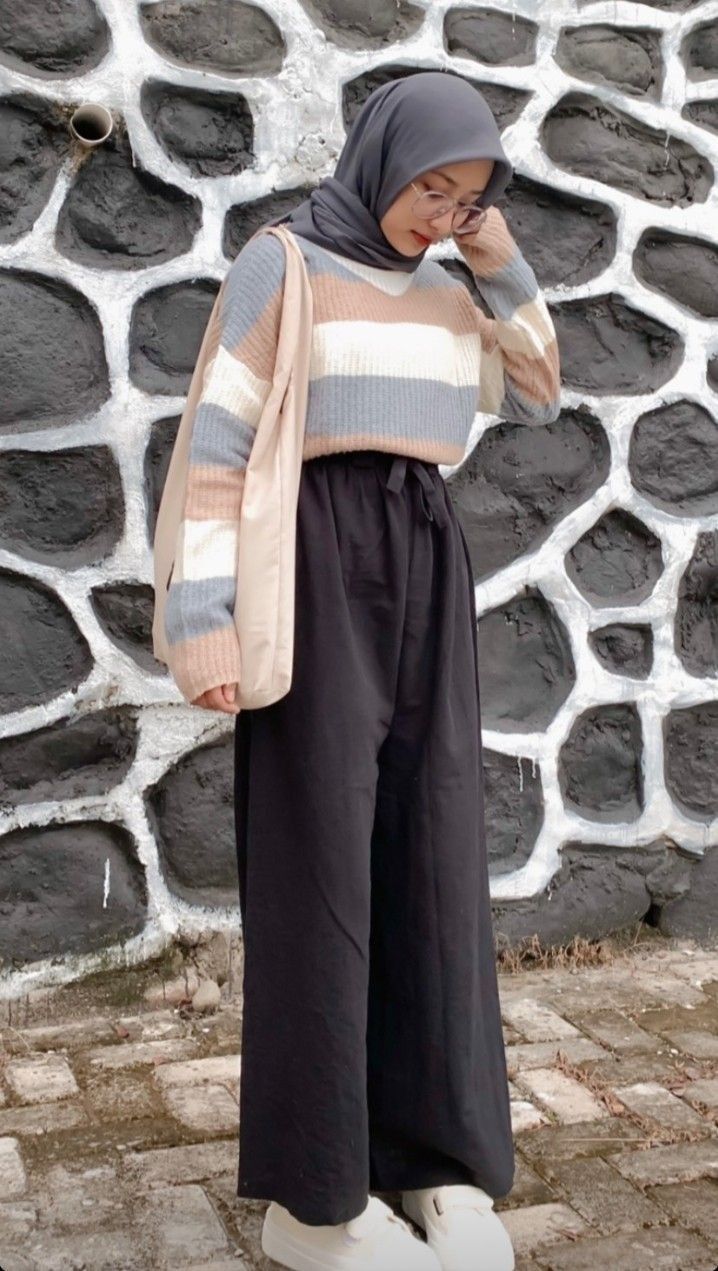 a woman standing in front of a stone wall talking on a cell phone while wearing black pants and a striped sweater