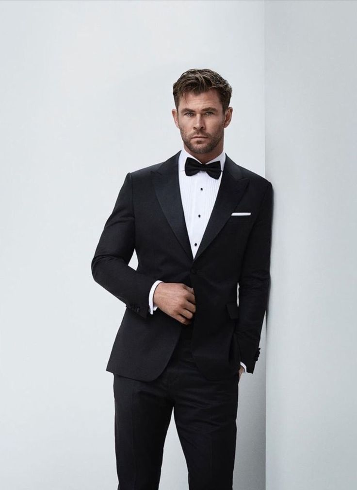 a man in a tuxedo leaning against a wall with his hands on his hips