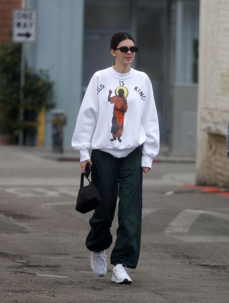 a woman walking down the street wearing a white sweatshirt and green pants