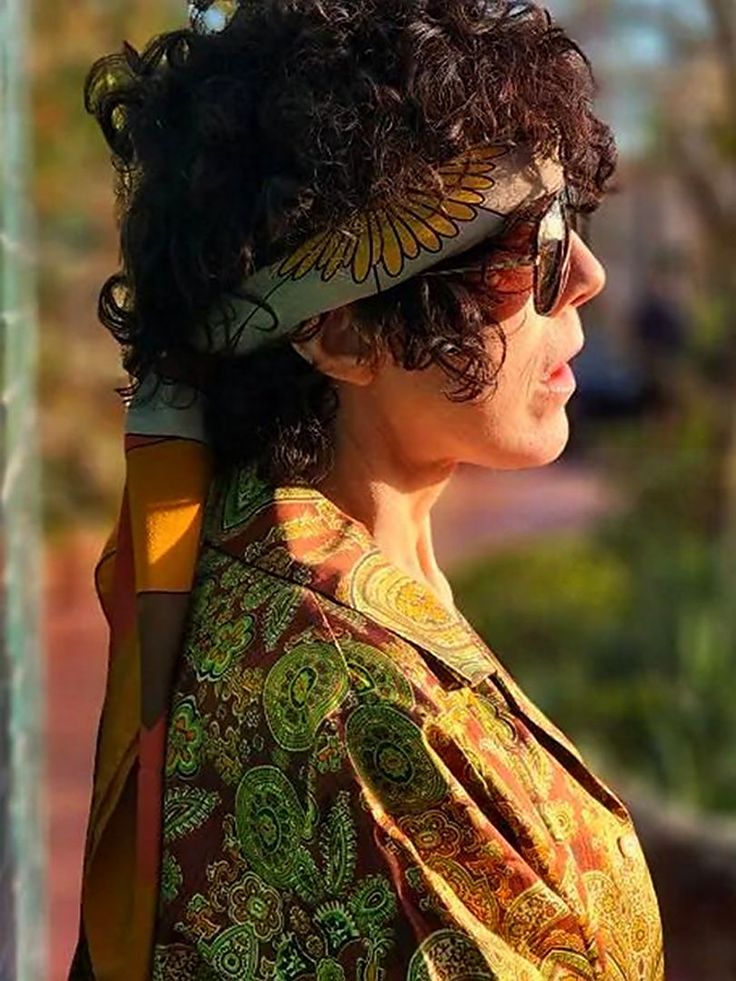 a woman with dark curly hair wearing sunglasses and a colorful shirt is looking off into the distance