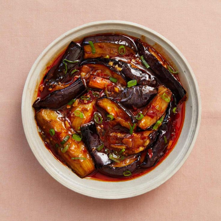 a white bowl filled with chicken and eggplant covered in sauce on top of a pink surface