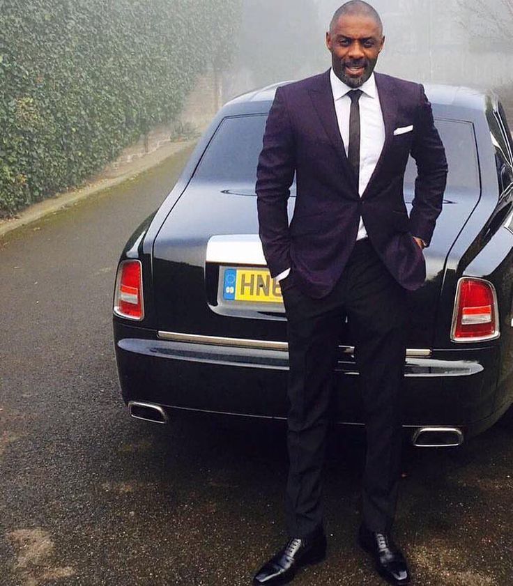 a man in a suit standing next to a car on a road with hedges behind him