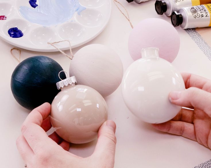 two hands are holding ornaments on a table with other crafting supplies and paint bottles