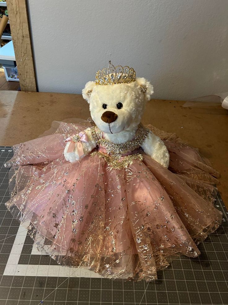 a teddy bear wearing a pink dress and a gold crown sitting on top of a table