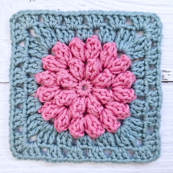 a crocheted square with a pink and blue flower on the center is sitting on a white wooden surface