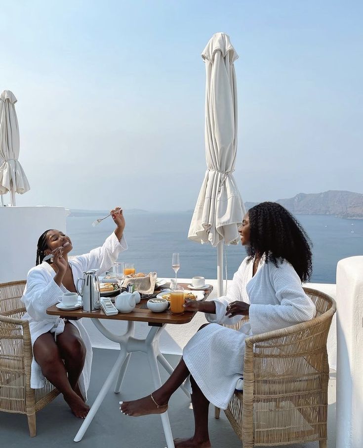 two women sitting at a table with food and drinks in front of an ocean view