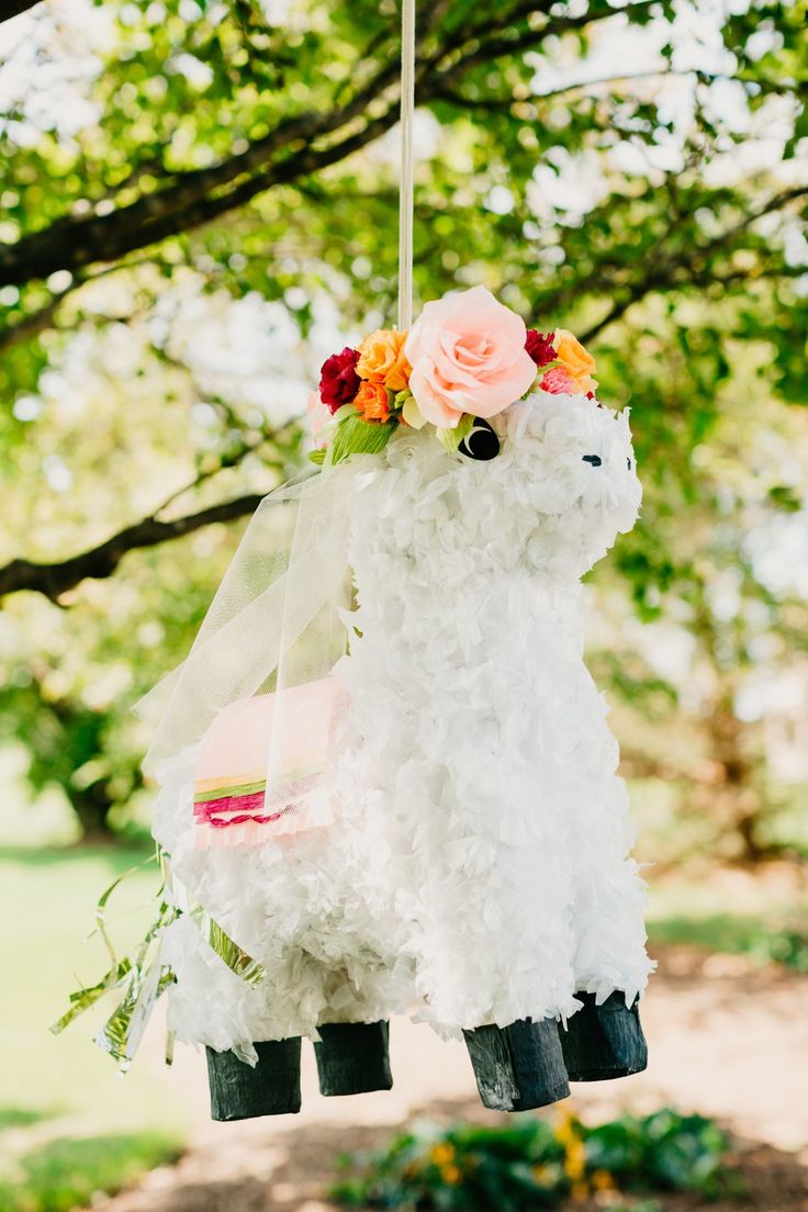 a white llama hanging from a tree with flowers on it's head and in the air