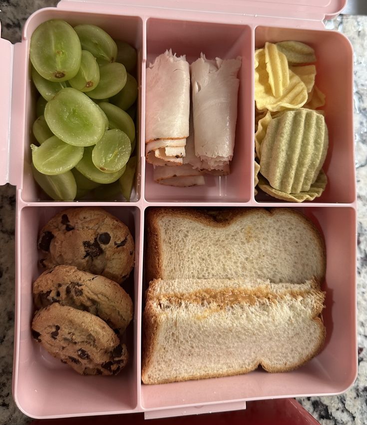 a pink lunch box filled with lots of food next to cookies and grapes on a table