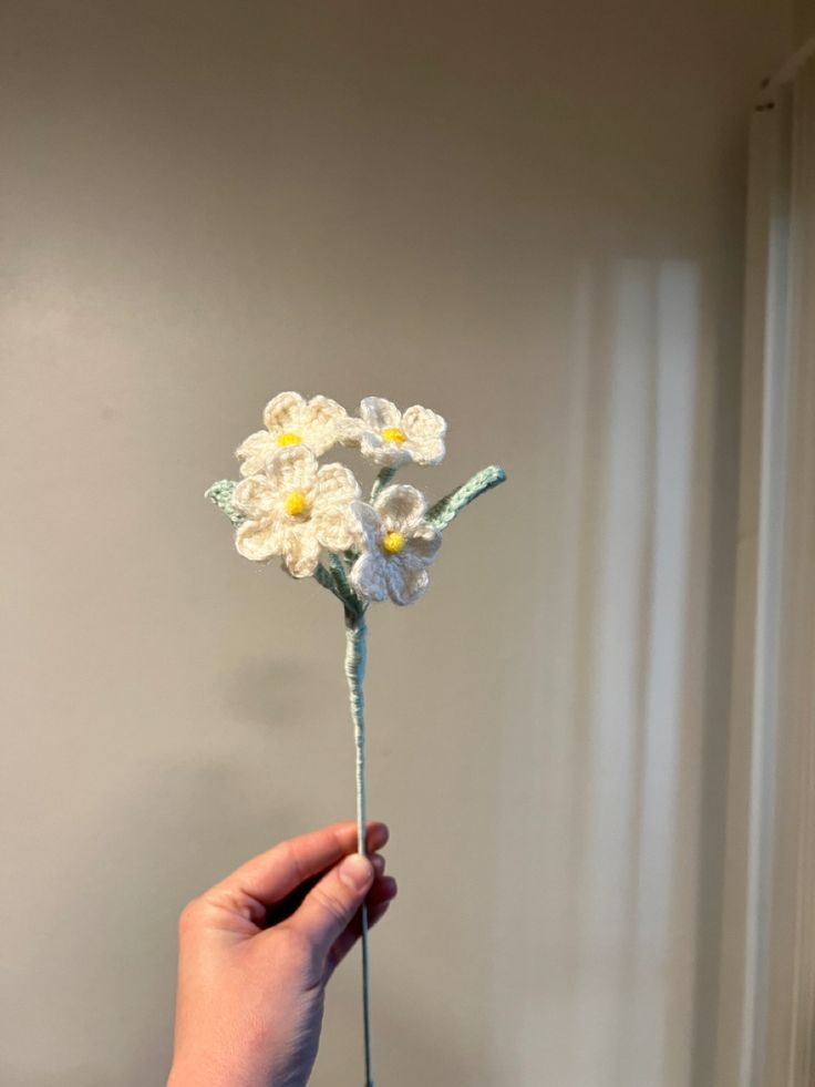 a hand holding a crocheted flower in front of a white wall