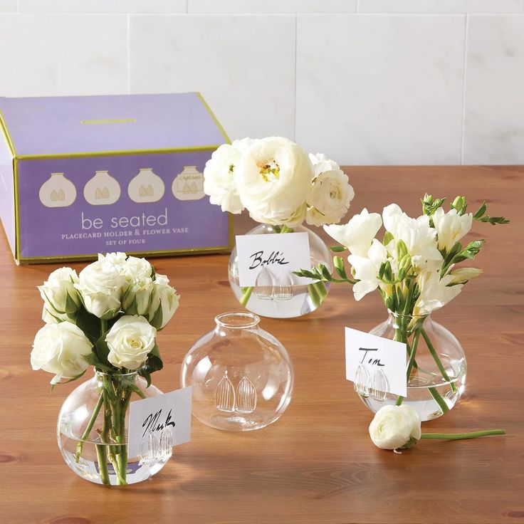 three clear vases filled with white flowers on top of a wooden table next to a purple box