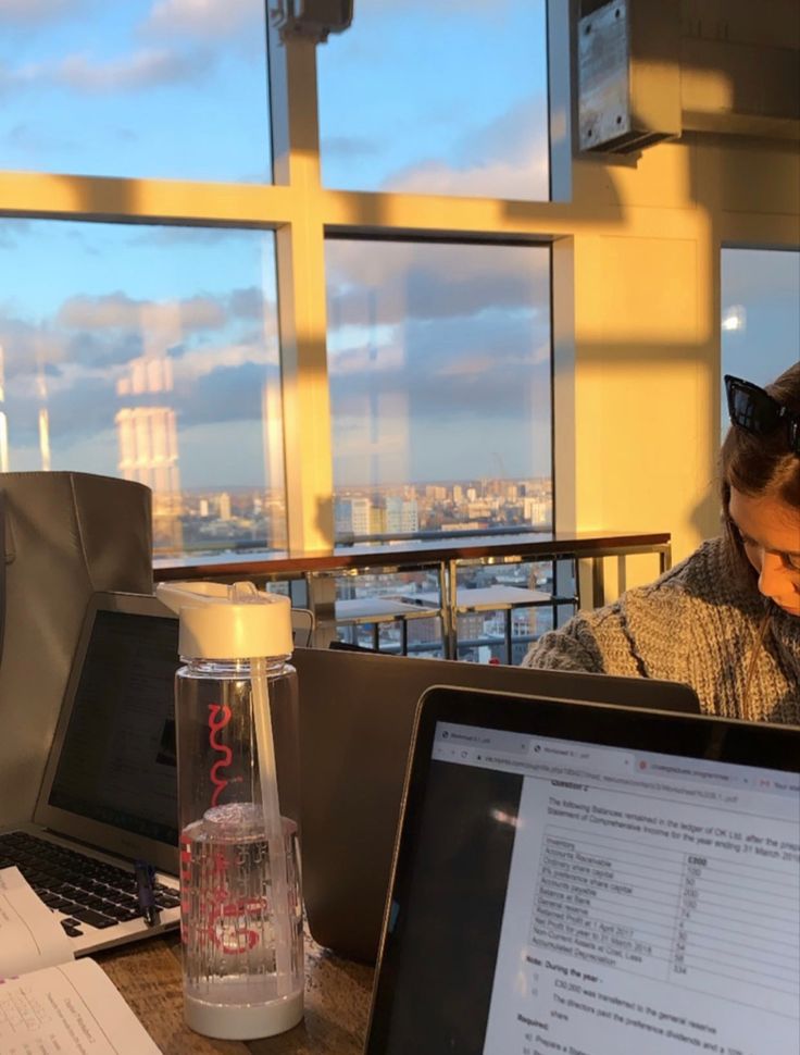 a man sitting at a desk with a laptop computer and monitor on it, in front of a large window