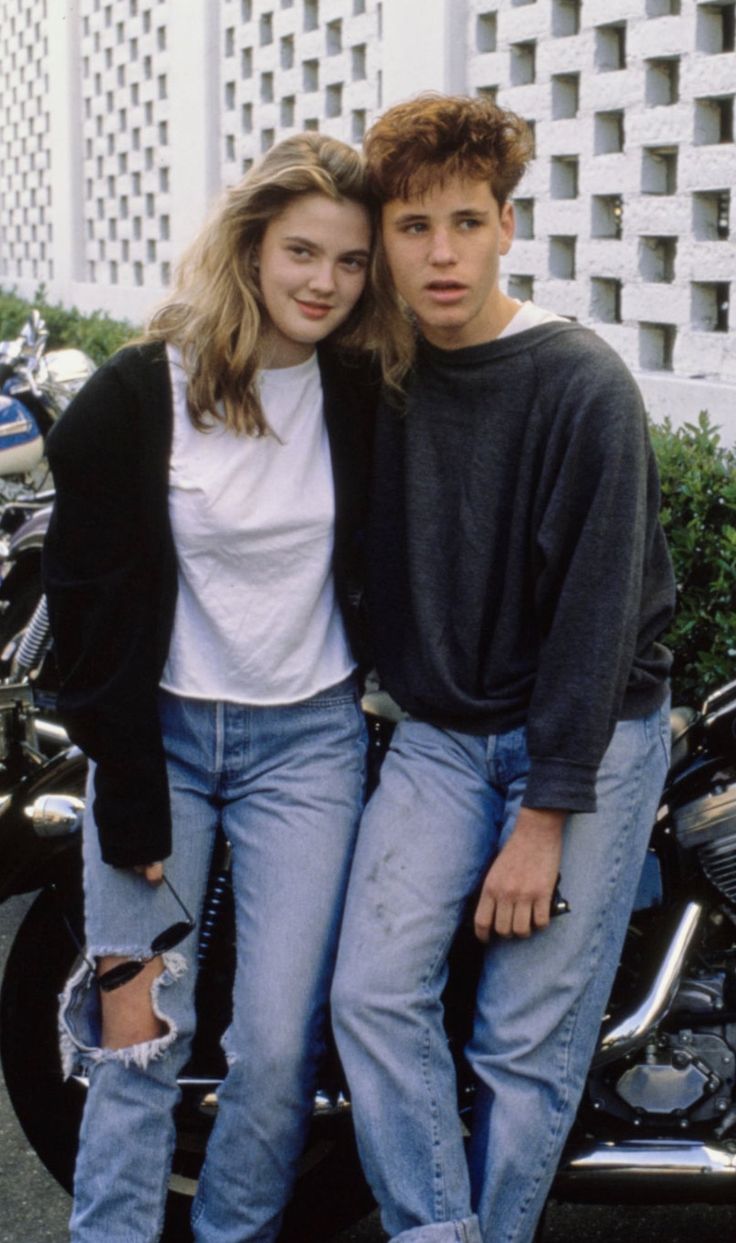 two people standing next to each other in front of a motorcycle with buildings behind them