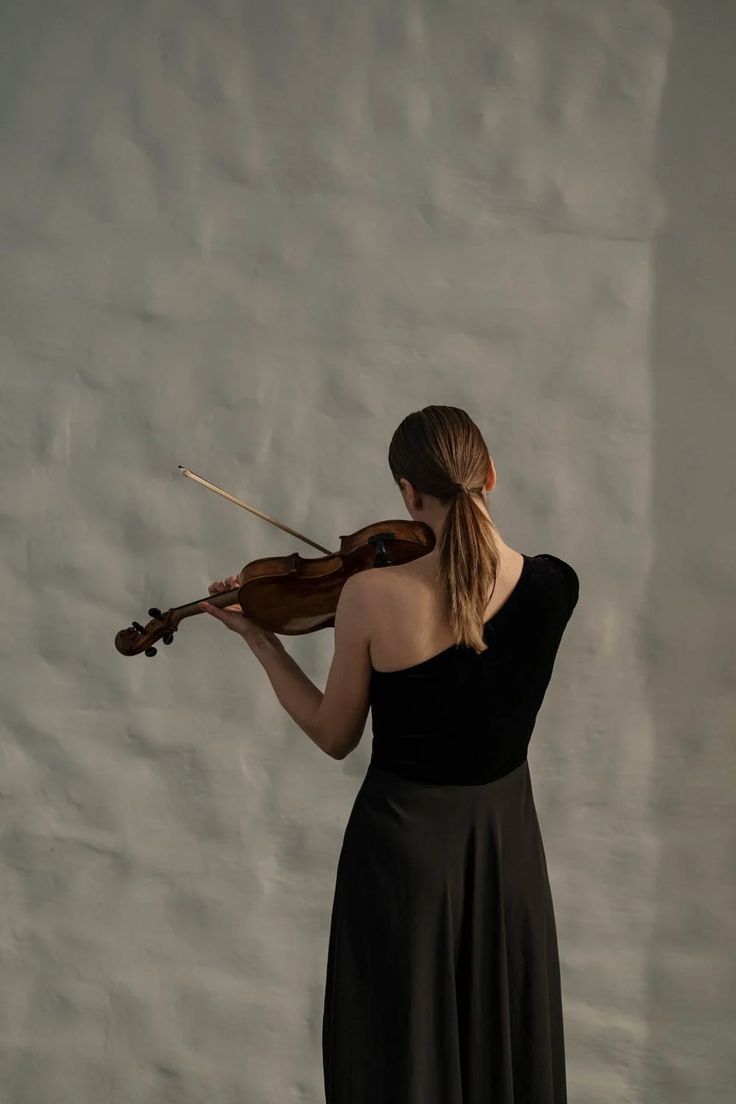 a woman in a black dress holding a violin and looking down at the ground while standing against a wall