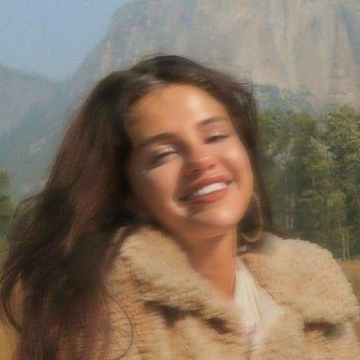 a woman with long hair smiling and wearing a fur coat in front of a mountain