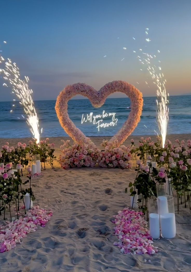 a heart shaped sign on the beach with flowers and candles