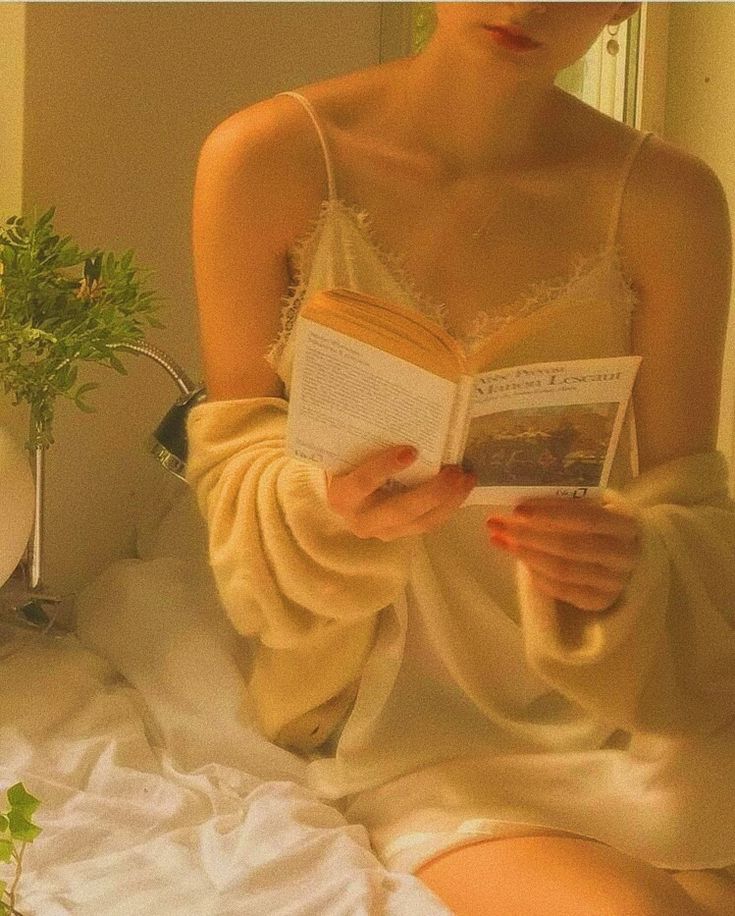 a woman sitting on a bed reading a book and holding a plant in her hand