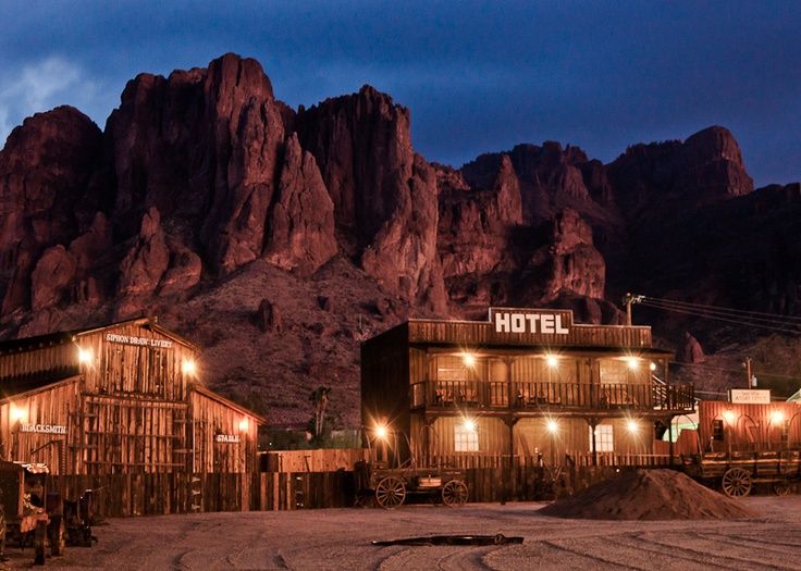 an old western town at night with mountains in the background and lit up hotel sign