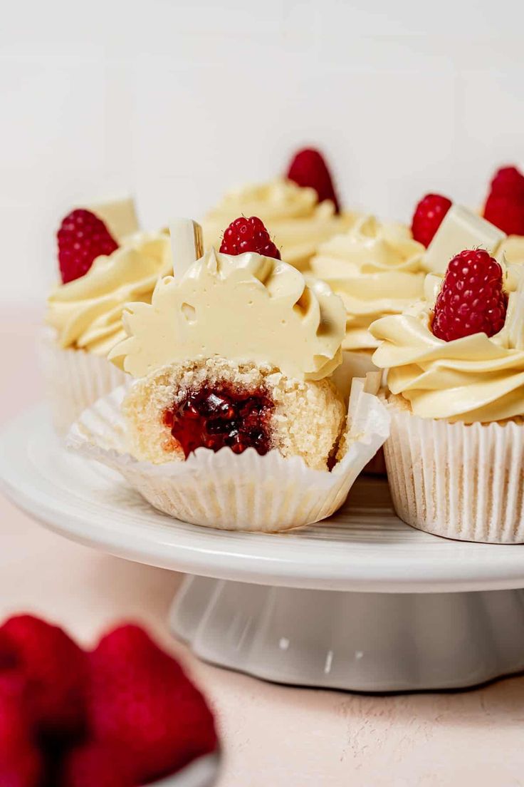 several cupcakes with white frosting and raspberries on a cake plate