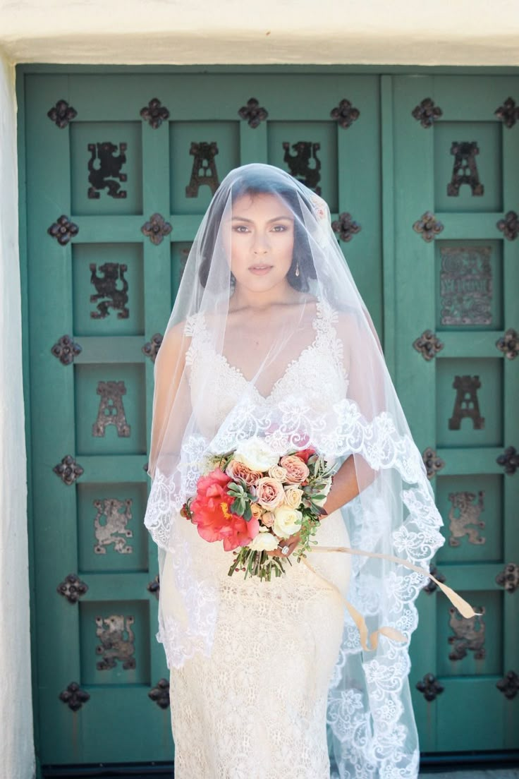 a woman standing in front of a green door wearing a veil and holding a bouquet