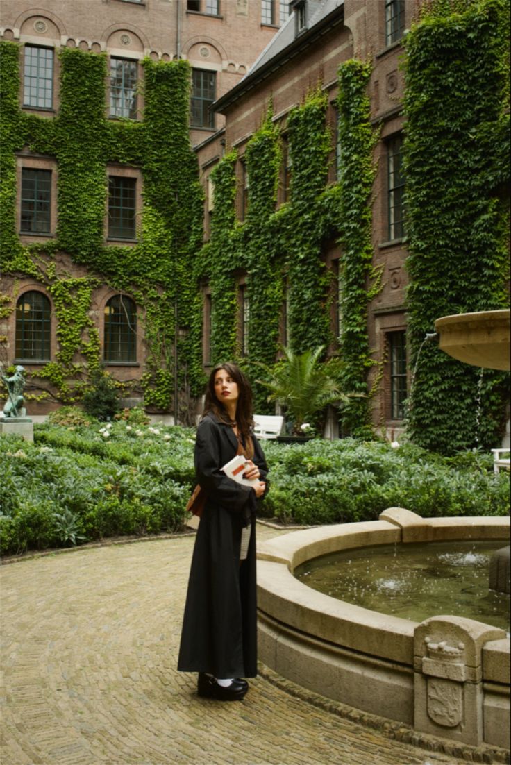 a woman standing in front of a fountain with ivy growing on the building behind her