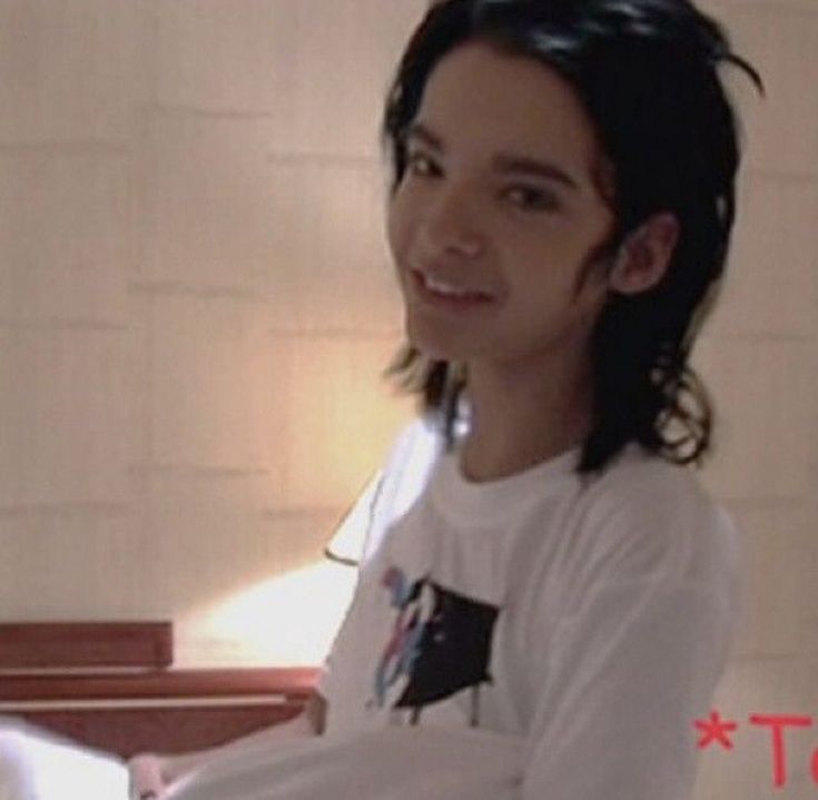 a young man with long black hair sitting in front of a piano and smiling at the camera