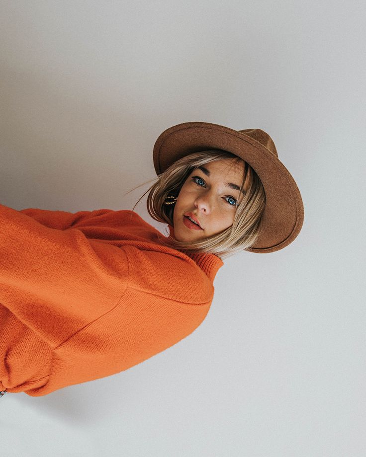 a woman in an orange sweater and brown hat poses for the camera with her hands on her hips