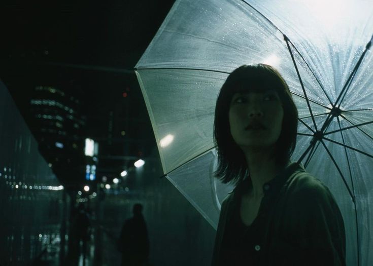 a woman standing in the rain holding an open umbrella at night with people walking by