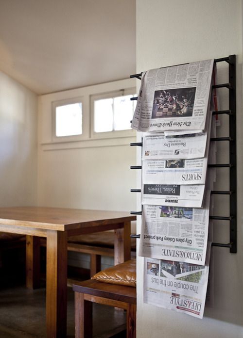 a wooden table sitting under a window next to a wall mounted newspaper rack with newspapers on it