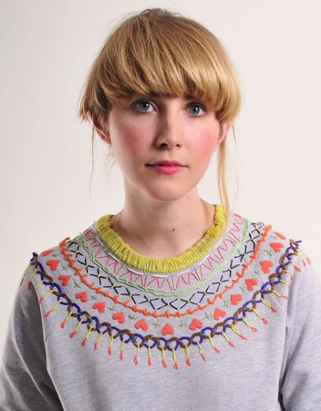 a woman with blonde hair and blue eyes wearing a gray sweater is standing in front of a white wall