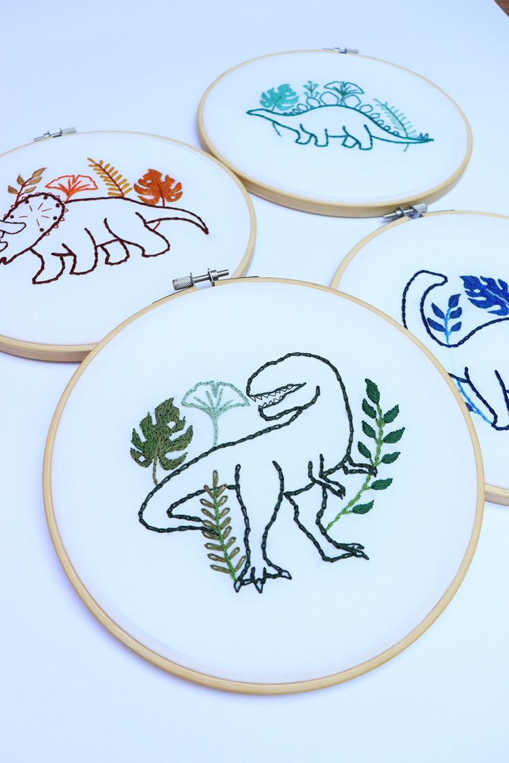 four embroidered dinosaurs are shown in different colors and sizes on white fabric with wooden hoop frames