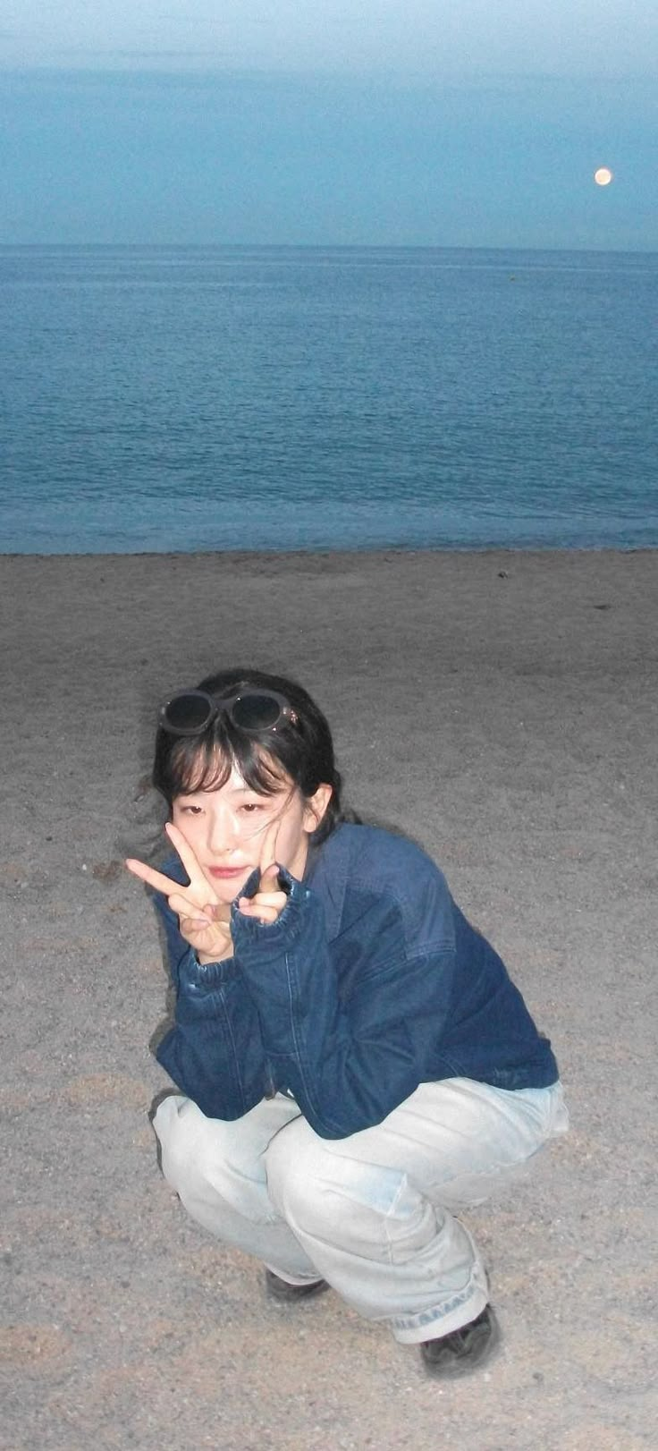 a woman kneeling down on top of a sandy beach next to the ocean holding a peace sign