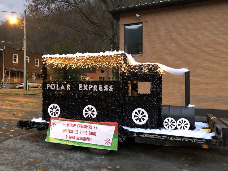 a small train is decorated with christmas lights and snow on the ground in front of a brick building