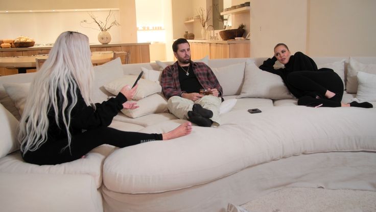 three people sitting on a white couch in a living room, one with long hair